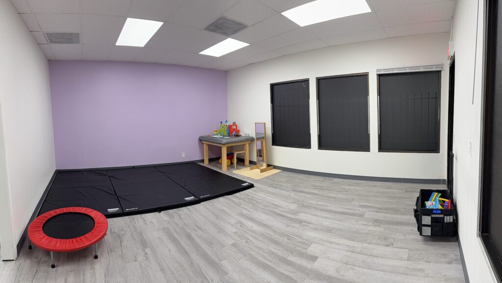 Purple-wall therapy room with black floor mats, a mini trampoline, a treatment table and windows at Sensory Bridge Pediatric Therapy.