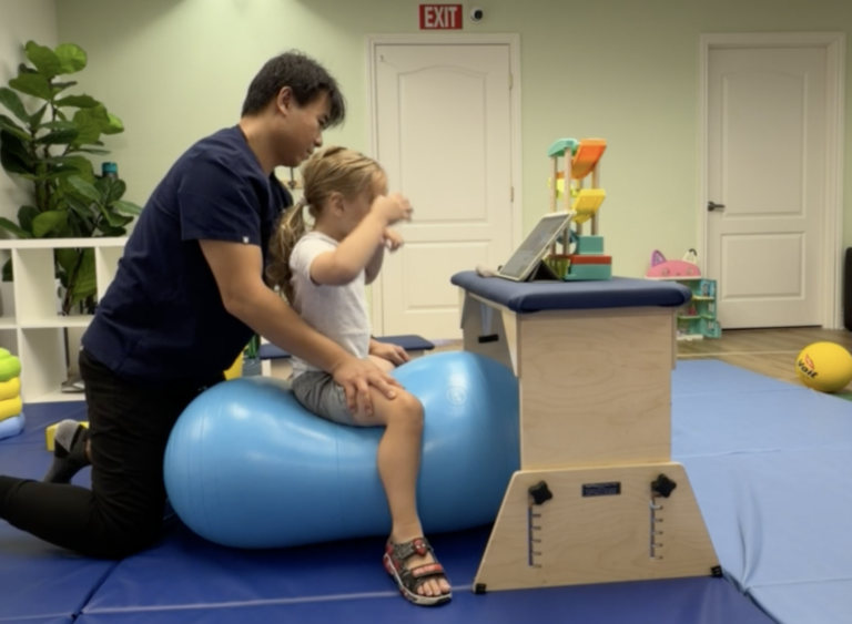 Therapist supporting a child seated on a blue peanut ball beside an activity table.