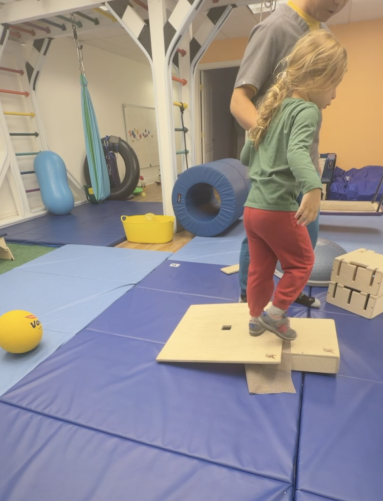Therapist supporting a child stepping onto a wooden DMI platform.
