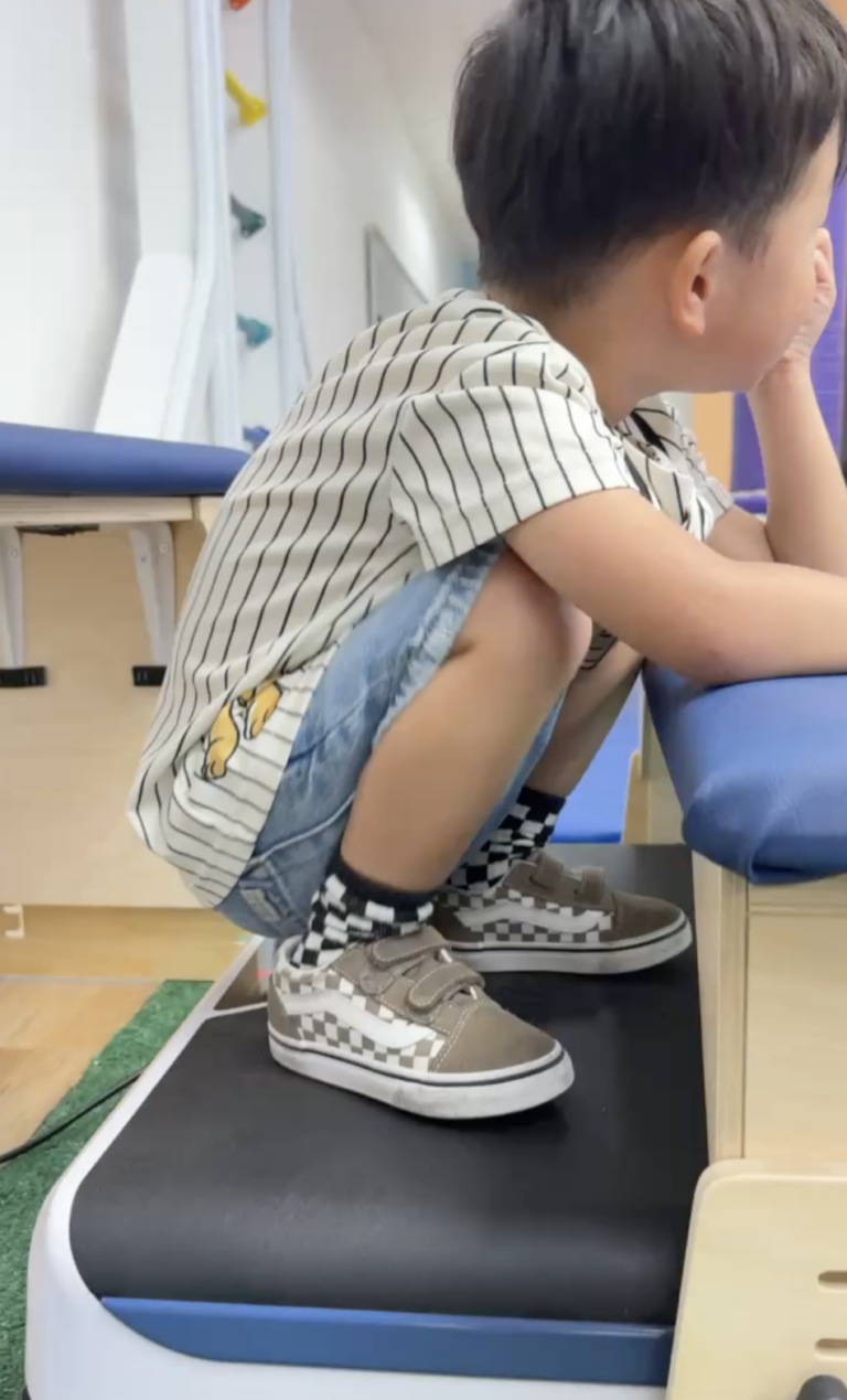 Child squatting on a vibration platform beside an activity table.