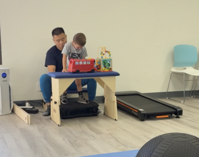 Therapist supporting a child standing on a vibration platform at an activity table.