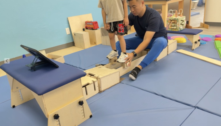 Therapist supporting a child's feet on wooden DMI beams beside an activity table.