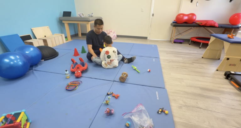 Therapist and child seated on floor mats with toys and colorful cones.
