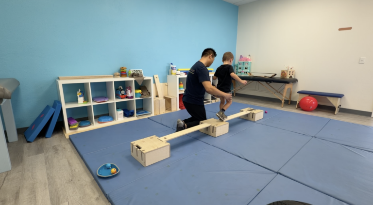 Therapist supports a child stepping along a wooden DMI balance beam.