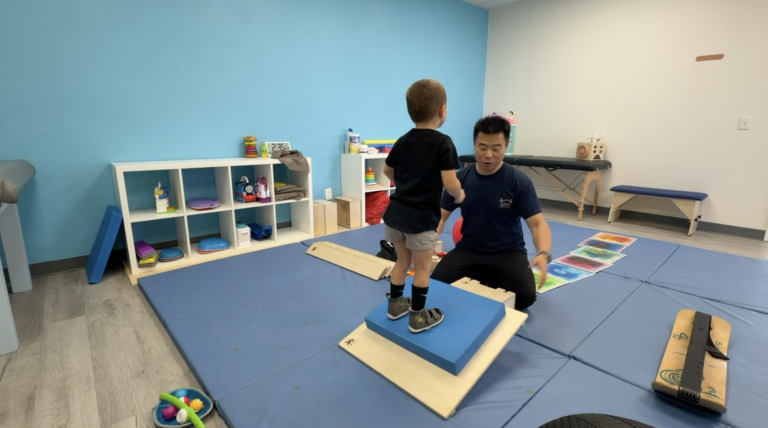 Therapist supervising a child standing on a foam pad placed on a wooden DMI platform.